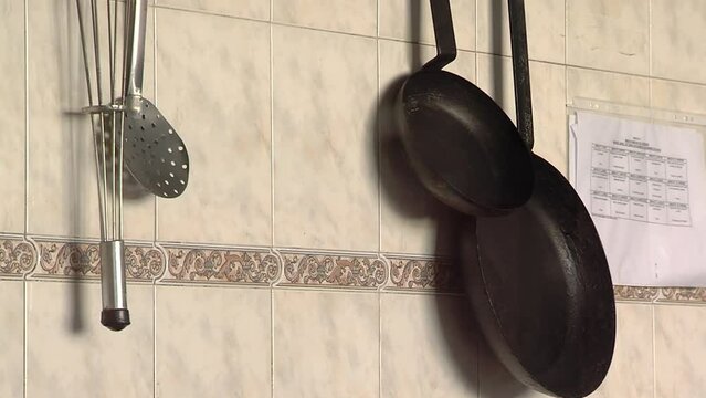 A Whisk, a Skimmer Spoon and Frying Pans Hanging from a Wall inside the Kitchen of a School Cafeteria. Close Up.