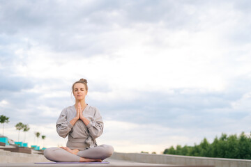 Fototapeta na wymiar Portrait of calm Caucasian young woman practicing yoga performing namaste pose with closed eyes outside in city park. Pretty serene female sitting lotus position on yoga mat outdoors alone.