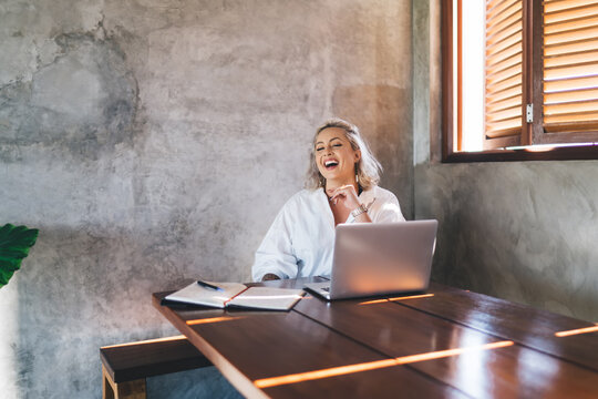 Joyful female software developer laughing and rejoicing during remote working indoors, millennial woman with laptop computer doing distance job and web design while smiling at desktop table