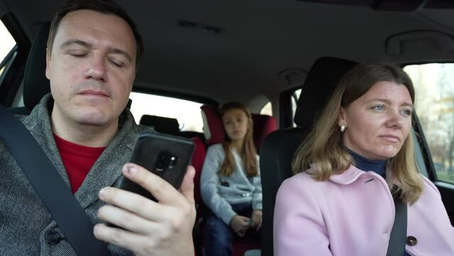 Girl driver drives car and man sitting in passenger seat, looking social media, texting messages using cell phone. Married couple with children on trip. Concept of travel, love and family