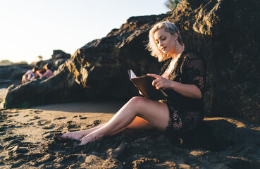 Beautiful female reading literature best seller sitting at sand and resting with hobby, charming Caucaisan woman in stylish wear holding education notepad analyzing book plot during vacations