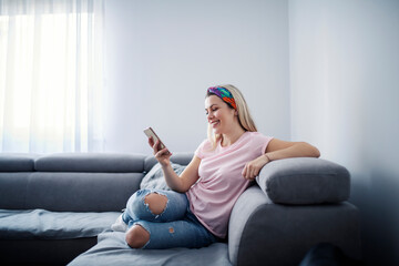 A happy woman sitting on couch at her comfortable home and texting a message on the phone.
