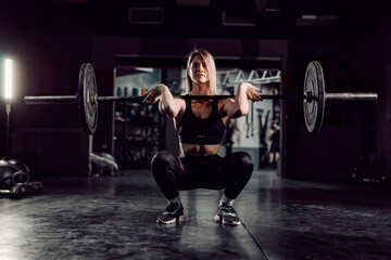 A female bodybuilder crouching at the gym and lifting barbell. 