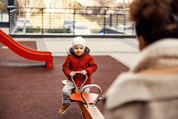 Play and games time. Happy little boy rocking on the seesaw with his mother at the playground.