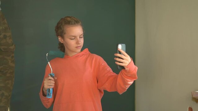 Cute girl with a paint roller in her hands takes a selfie while her father paints the walls in her room. Social media and teenagers concept
