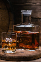 Whiskey with ice, vertical format. Decanter of old whiskey and whiskey in glass of ice against backdrop of old barrel of full alcoholic beverage on wooden background in old basement.