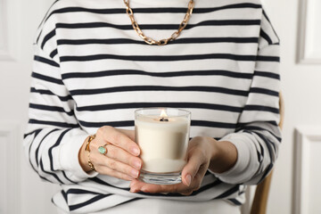 Woman with stylish jewelry holding burning soy candle on light background, closeup