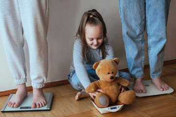 The family is weighed at home, two young women stand on the scales, a little girl copies them and weighs a toy teddy bear on a kitchen scale.