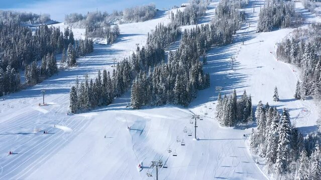Aerial view of ski resort with people skiing and snowboarding down the hill. Flying over the ski lift, ski or snowboard track on white snow surrounded by trees in winter season