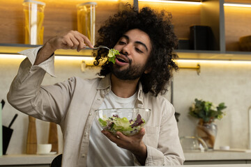 Egyptian man with dark kinky hair enjoys eating salad with lettuce from transparent bowl at table...