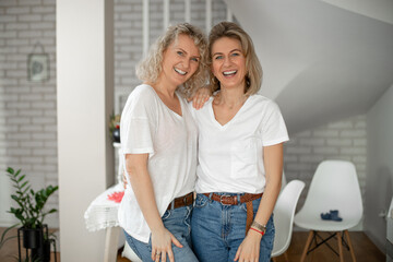 Portrait of two blond smiling, gleeful women mother and daughter hugging in kitchen enjoy and hangout together.Side view