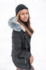 beautiful young African model woman in fashionable clothes with a stylish winter jacket and hat walks outdoors with snow. Fashion pretty girl on a white background