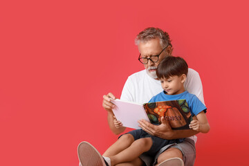 Senior man reading book to his little grandson on red background