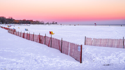 A snow covered and mostly empty beach  with snow snow fences in the foreground, sky tinged with pink and red.  Shot in Toronto in January.