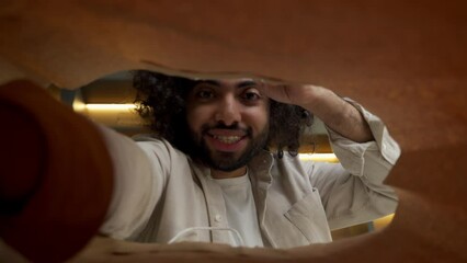 Cheerful Muslim man with kinky hair opens paper bag to take out products delivered from grocery store in kitchen close view from inside