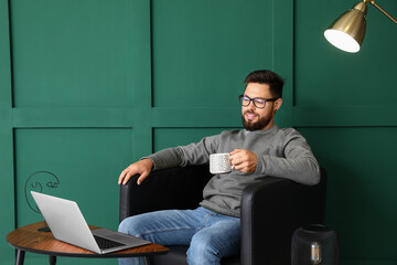 Handsome bearded man with cup of hot beverage sitting in leather armchair near green wall