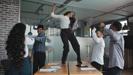 Joyful diverse employees celebrating victory project completion female leader smiling happily dancing on table multiracial colleagues at office party happy friendly work group of having fun together