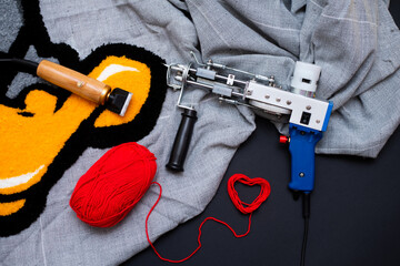 The tufting gun lies on a gray burlap with a bright rug, a wooden lint clipper and red yarn in the...