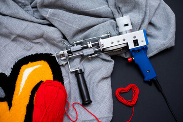 The tufting gun lies on a gray burlap with a bright rug, a wooden lint clipper and red yarn in the...