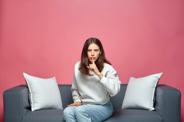 Shh, don't speak. Serious modern girl show silence gesture, ask to stay quiet, sitting on sofa on pink studio background