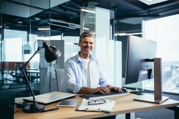 Obraz premium Portrait of a successful businessman, man rejoices and smiles, works at the computer, gray-haired and experienced looks at the camera