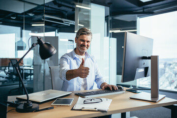 Portrait of a successful businessman, man rejoices and smiles, works at the computer, gray-haired and experienced looks at the camera