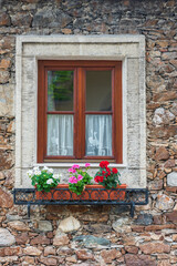 Fototapeta na wymiar A wooden, brown window and a fragment of a stone wall of the building. Curtains hanging in the window, colorful flowers on the windowsill.