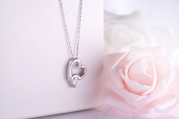 Diamond heart necklace hanging on white jewelry box soft roses in background - obrazy, fototapety, plakaty