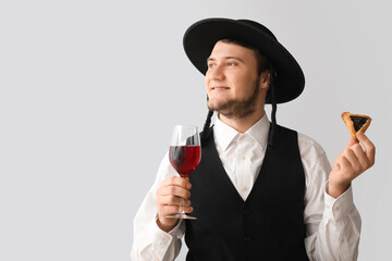 Young Jewish man with tasty Hamantaschen for Purim holiday and glass of wine on grey background