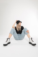 Fashionable pretty young model woman with Panama hat in stylish clothes with blue jeans and white leather shoes sits and poses on white background in studio. Beautiful blonde girl with summer outwear