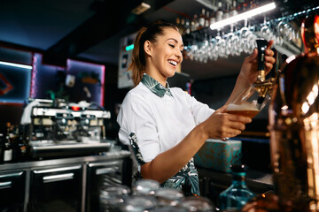 Happy female barista pours beer from a beer tap while working in pub.