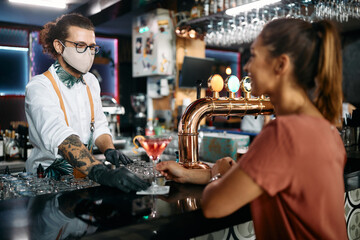 Professional barista wears protective face mask while serving cocktail to female customer at bar counter.