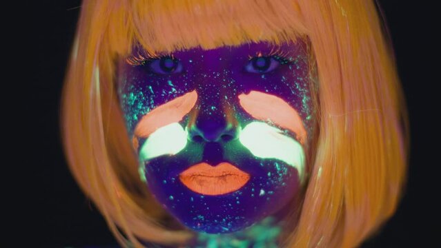 Neon paint. Young woman with neon body art and orange wig painting fluorescent green die on her face, dancing to camera