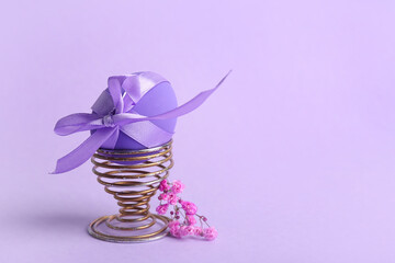 Holder with beautiful Easter egg and flowers on purple background
