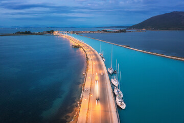 Aerial view of beautiful road near sea canal at night in summer in Lefkada, Greece. Top view of road, blurred cars, boat and yachts, city lights, azure water, mountain and cloudy sky at dusk. Travel  