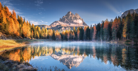 Lake with reflection of mountains at sunrise in autumn in Dolomites, Italy. Landscape with Antorno lake, blue fog over the water, trees with orange leaves and high rocks in fall. Colorful forest 
