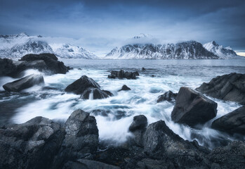 Beach with stones in blurred waves, blue sky with low clouds and snowy mountains at dusk in winter. Sea coast in Lofoten islands, Norway. Dramatic landscape with sea, rocks in fog. Moody scenery