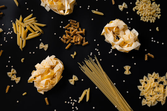 Different types of uncooked dried pasta on black background. Raw pappardelle, penne, spaghetti, farfalle and fusilli. Italian pasta cooking concept