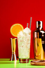 Tom Collins alcoholic cocktail with dry gin, syrup, lemon juice, soda, orange and ice. Trendy summer red green background, bar tools, copy space