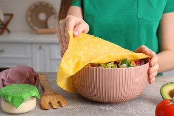 Woman covering bowl of fresh salad with beeswax food wrap at light grey table in kitchen, closeup - obrazy, fototapety, plakaty