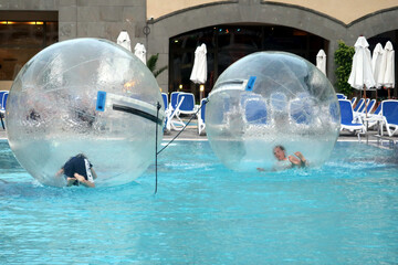 Children have fun inside big plastic balloons on the water of swimming pool. Children inside big...