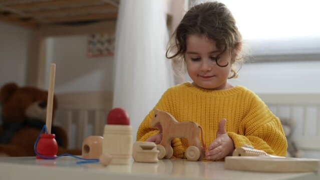 Adorable little girl with curly hair plays with with wooden toys at the table in children's room. Child has fun playing in her bedroom with eco toys. Education and child development. Lifestyle.