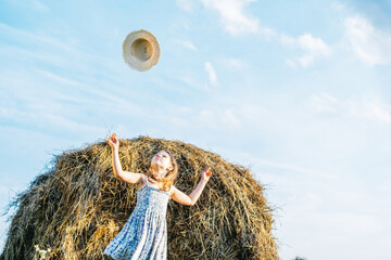 Portrait of little girl in blue dress catching hat, standing near haystack in field. Light sunny...