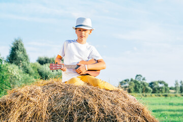 Naklejka na ściany i meble Portrait of barefoot boy in hat on haystack in field. Playing small guitar, ukulele. Light sunny day. Cheerful and music concept. Outdoor activity. Trees on background, Countryside