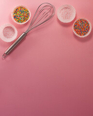 Cupcake liners with colored sugar inside whisk from above flat lay top view. Confectionery cooking concept with copy space on bright pink paper background. National cooking day