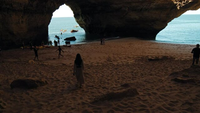 Beautiful girl in a dress walking, running in slow-motion in a rocky sandy cave. A fashion tourist woman barefoot on sand exploring Algar de Benagil Cave attraction in Algarve, Portugal.