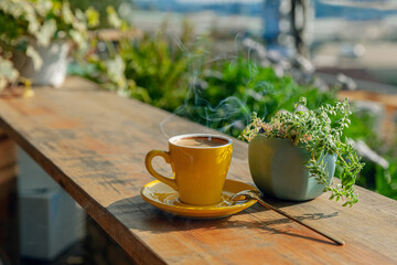 A cup of hot coffee is next to a potted plant on a wooden table, in the afternoon sunlight