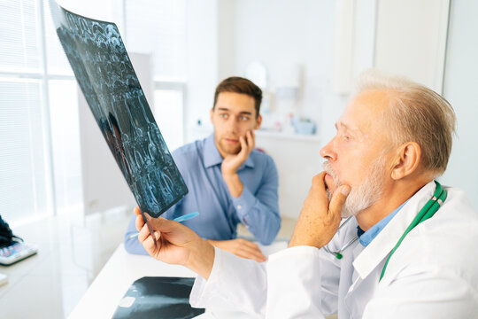 Thoughtful mature adult male physician consult frustrated young man patient giving bad news explaining results of MRI image. Unhappy scared young man listening to bad news sitting in doctor office.
