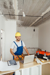 Happy repairman worker in uniform wearing tool belt and hardhat holding hammer and smiling at camera while standing indoors
