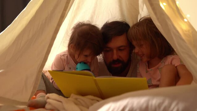 Happy dad and little daughters reading a book in a teepee tent decorated with lights in the darkroom in the evening. Cute kids at home with their father.